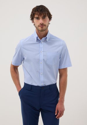 Hombre con camisa de manga corta de cuadros azul claro metida en pantalones azul oscuro, de pie con una mano en el bolsillo.