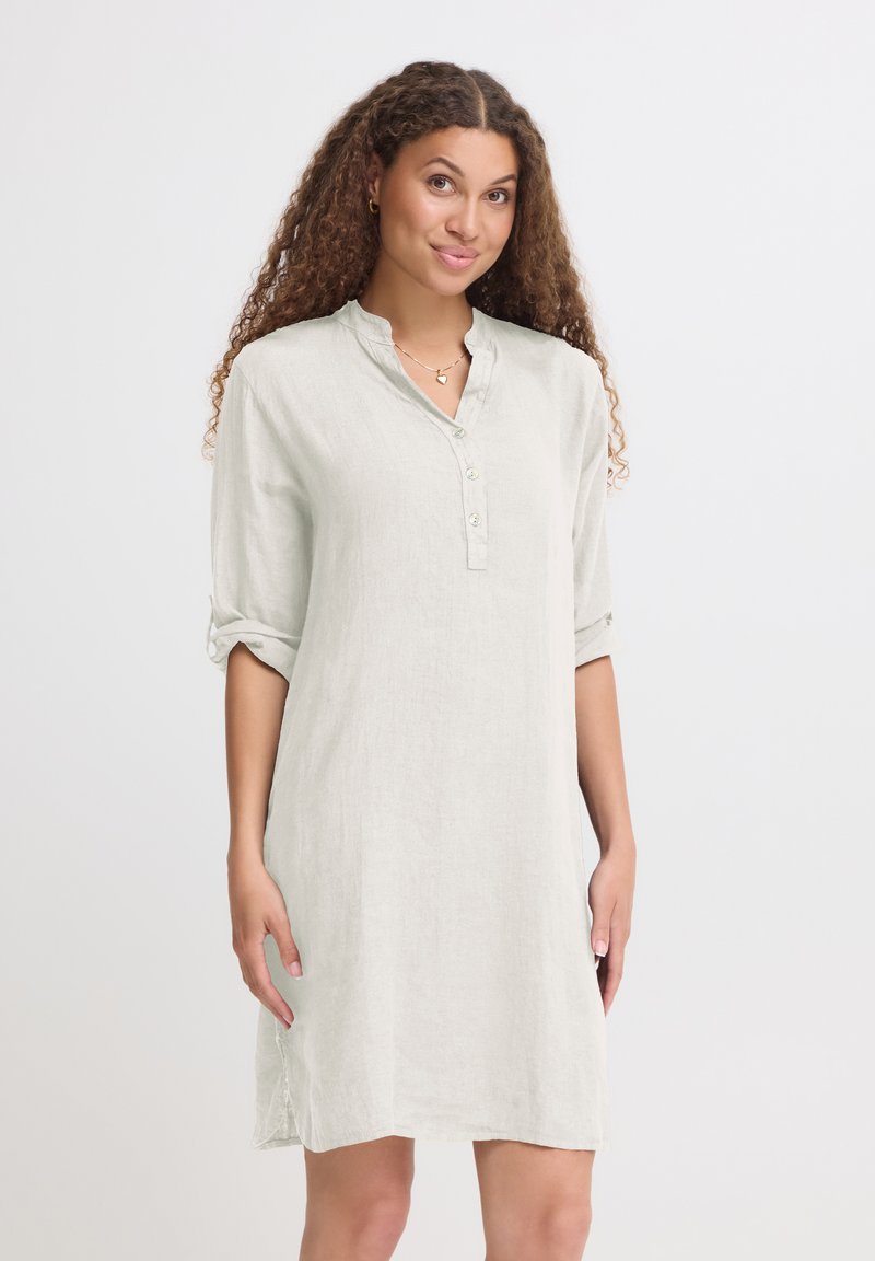 Femme aux cheveux bouclés portant une robe beige clair à manches longues avec manches retroussées et col boutonné, debout devant un fond uni.