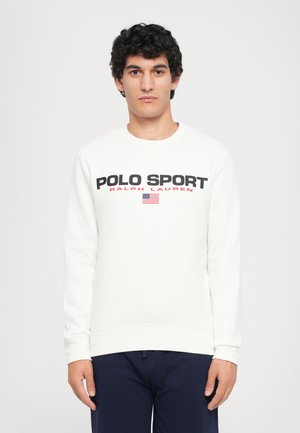 Ung mann med krøllete hår iført en hvit "Polo Sport Ralph Lauren" sweatshirt og mørke bukser, stående foran en enkel hvit bakgrunn.