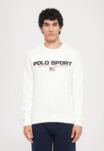 Młody mężczyzna z kręconymi włosami, ubrany w biały sweatshirt "Polo Sport Ralph Lauren" i ciemne spodnie, stojący na jednolitym białym tle.