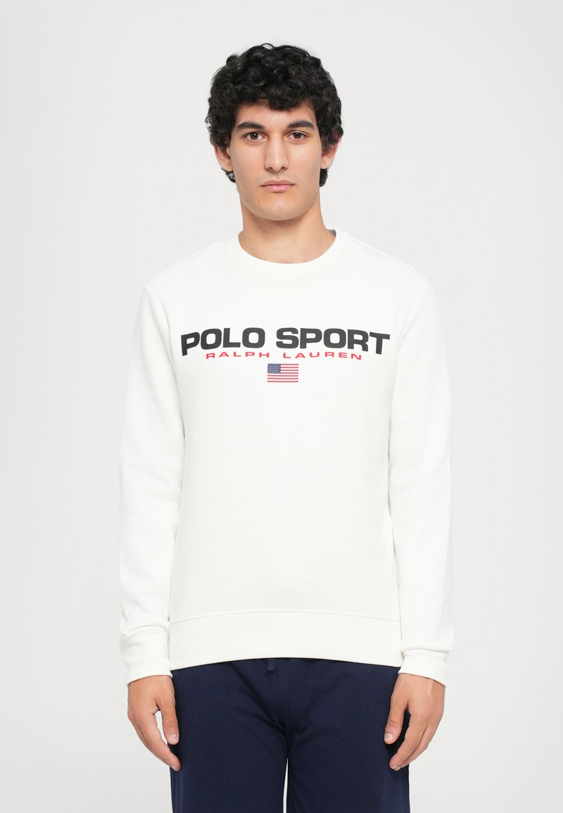 Tânăr cu păr creț, purtând un pulover alb "Polo Sport Ralph Lauren" și pantaloni închiși la culoare, stând pe un fundal alb simplu.
