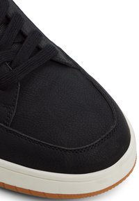 Zapatilla de deporte negra de cuero sintético con superficie texturizada, media suela blanca en contraste y suela de goma marrón. Los cordones son negros y están pasados por las ojetes.