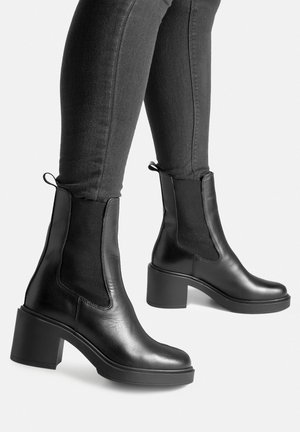 Czarne skórzane botki za kostkę z elastycznymi panelami po bokach, masywnym obcasem i gładką fakturą, zestawione z dopasowanymi ciemnymi jeansami.