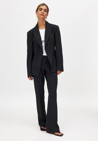 Črn prirejen suknjič z enim gumbom, povezan z širokimi hlačami. Tkanina ima gladko teksturo, oblačilo pa je ujemljeno z belo majico.