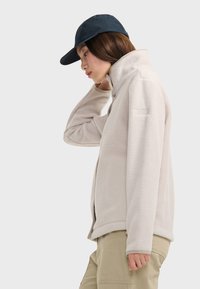 Chaqueta de forro polar beige con cuello alto, cierre de cremallera y bolsillos laterales. Textura suave y detalles en contraste en los puños y el dobladillo.