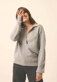 Grauer, gerippter Pullover mit weitem Kragen und lockerer Passform. Lange Ärmel und tiefes V-Ausschnitt-Design. Kombiniert mit dunklen Hosen.