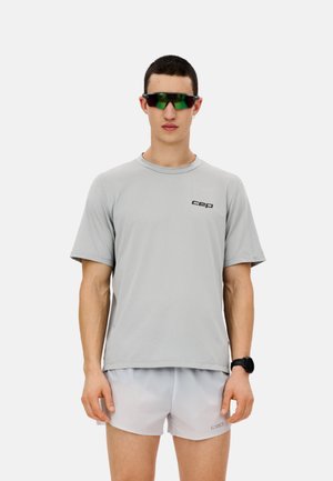 Mann trägt graues Sportshirt und Shorts, grün getönte Sportsonnenbrille und schwarze Armbanduhr, steht vor weißem Hintergrund.