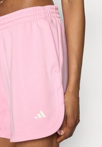 Világos rózsaszín sportnadrág puha anyagból, rugalmas derékpánttal, oldalzsebekkel és fehér Adidas logóval a szegélyen.