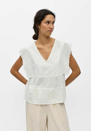 Femme aux cheveux foncés coiffée en chignon bas porte un haut sans manches en dentelle œillet blanche avec des bords festonnés et un pantalon beige, regardant sur le côté.