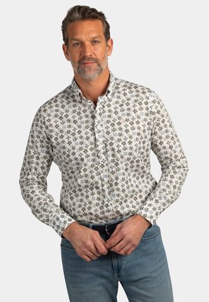 Man wearing light grey floral button-up shirt and blue jeans against plain background, adjusting belt with both hands.