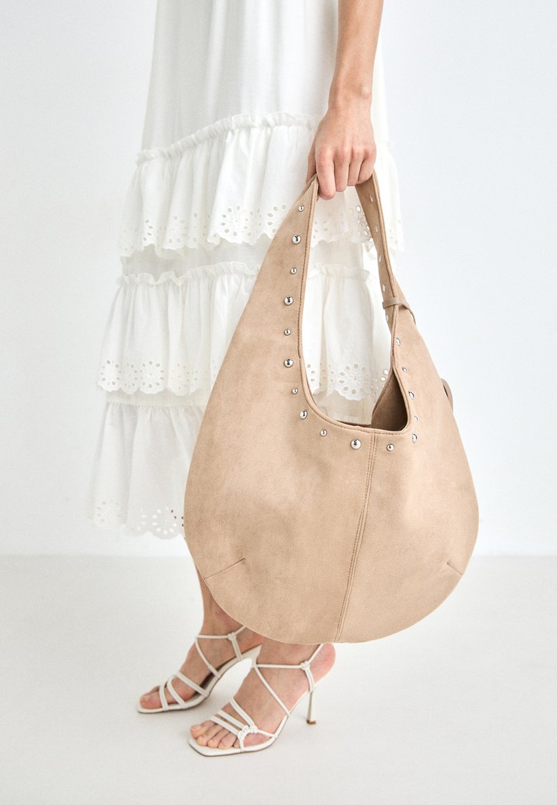 Bolso hobo de ante color canela con tachuelas plateadas a lo largo del borde superior, con una forma redondeada y una sola correa, sostenido por una persona.