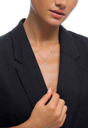Silver cross necklace on a thin chain worn with a black blazer. The necklace features a simple, polished design and hangs just below the collar.