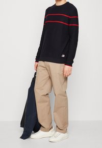 Pull marin avec deux rayures rouges sur la poitrine, associé à un pantalon beige et des baskets blanches. Tient une veste marine.