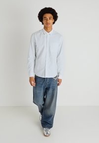 Ανοιχτό μπλε ριγέ πουκάμισο με τσέπη στο στήθος, συνδυασμένο με φαρδιά ξεθωριασμένα μπλε τζιν και ασημί αθλητικά παπούτσια.