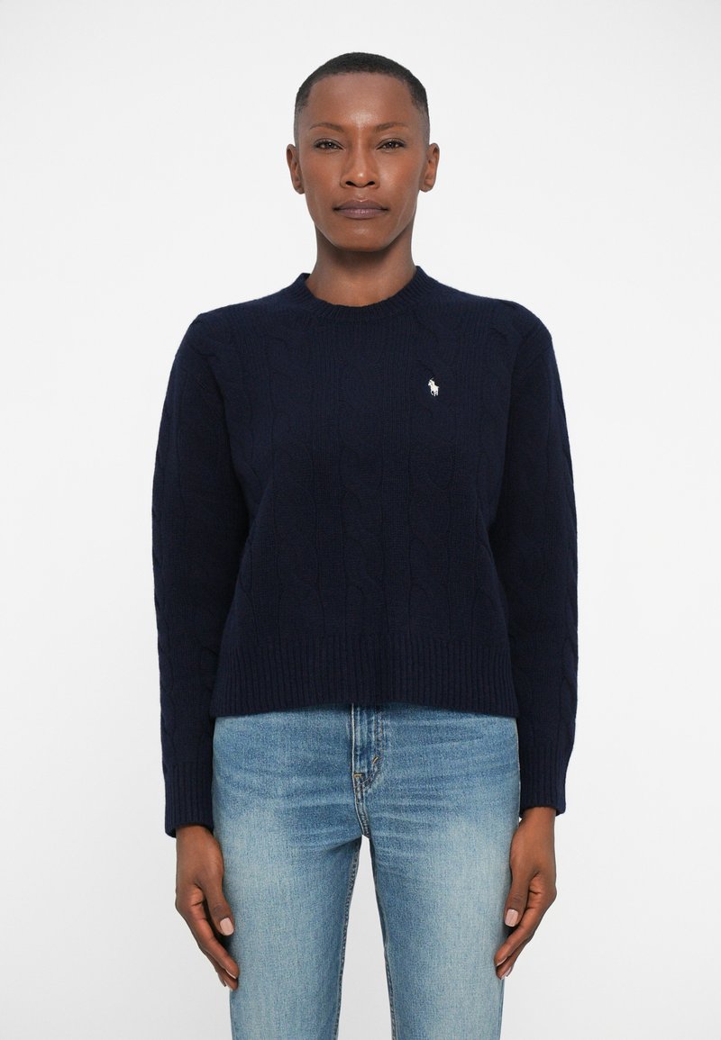 Navy cable-knit sweater with ribbed hem and cuffs, featuring a small white logo on the chest. Worn with light wash denim jeans.