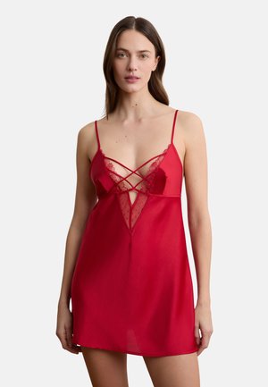 Kobieta ubrana w czerwoną satynową sukienkę na ramiączkach z koronkowymi dodatkami i krzyżującymi się paskami na piersiach, stojąca na tle jasnego, jednolitego tła.