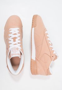 Ljust rosa sneakers i mjukt mockamaterial, vita snören, perforerat tåparti och en vit sula. Har Reebok-logga och klassisk design.