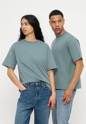 Young woman and man wearing matching muted green t-shirts and blue jeans, standing side by side and looking in opposite directions.