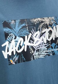 Blaues Baumwoll-T-Shirt mit einem rechteckigen Grafikdesign, das tropische Blätter und einen Vogel in Schwarz-, Blau- und Weißtönen zeigt; großer Text lautet "JACKSON."