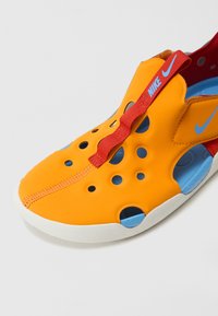 Sneaker en caoutchouc orange avec design perforé, intérieur bleu, semelle blanche et sangle rouge. Logo Nike en bleu sur le dessus.