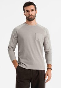 Uomo con barba che indossa una camicia a maniche lunghe grigio chiaro con taschino e pantaloni scuri, in piedi con una mano in tasca.