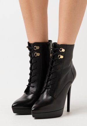 Zwarte leren enkelboots met een hoge hak, spitsen, vetersluiting aan de voorkant en accenten in goudkleurig beslag. Ritssluiting aan de zijkant.
