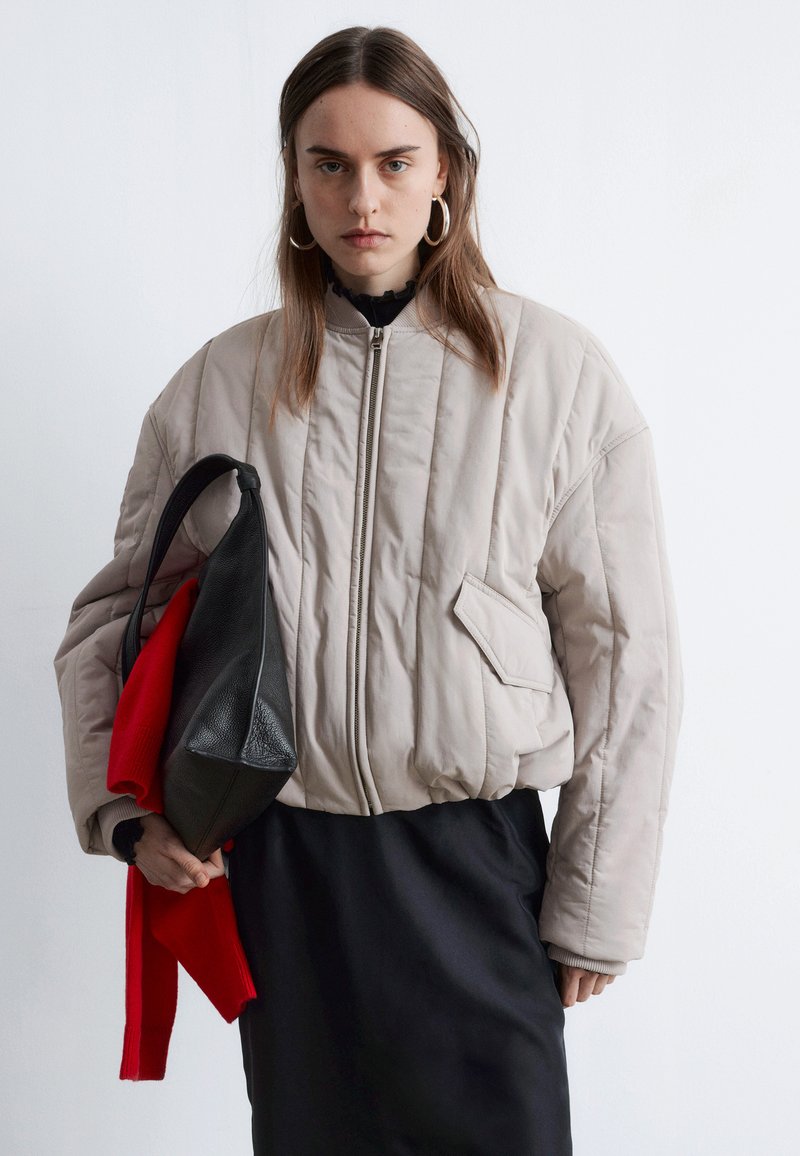 Doudoune beige clair avec un motif matelassé, fermeture éclair et poche latérale. Associée à une jupe noire et tenant un sac rouge et noir.