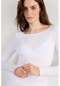 Jonge vrouw met lichte make-up en los bruin haar, gekleed in een doorschijnend wit shirt met lange mouwen, staand tegen een witte muur.