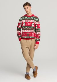 Maglione di Natale lavorato a maglia rosso e verde con pupazzi di neve e motivi; abbinato a pantaloni di corduroy beige e scarpe marroni.