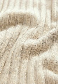 Beige Strickstoff mit geripptem Muster. Leichter Schimmer von eingewebten metallischen Fäden, die subtile Tiefe und Dimension erzeugen.