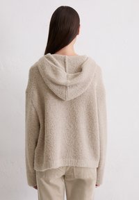 Felpa beige con cappuccio in pile, dal tessuto morbido, taglio oversize e cappuccio profondo. Presenta una cucitura centrale che scorre lungo la schiena. Abbinata a pantaloni beige.
