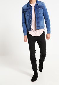 Chaqueta de mezclilla azul con dos bolsillos en el pecho, llevada sobre una camisa rosa claro, combinada con jeans negros desgastados y botas de tobillo negras.