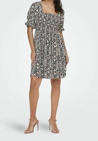 Robe à motif noir et blanc avec un décolleté carré, des manches bouffantes et une taille froncé, tombe au-dessus du genou. Portée avec des talons nus à lanières.