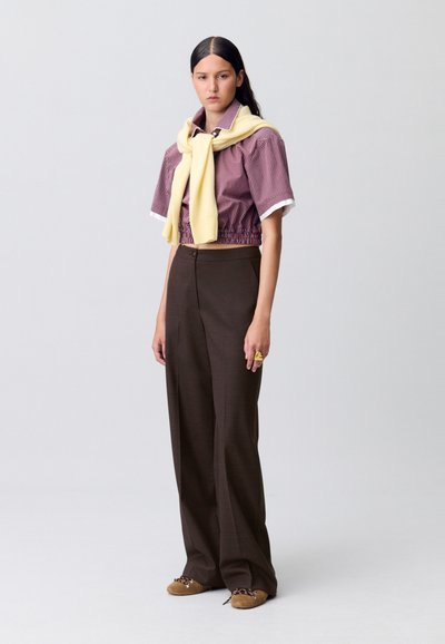 Pantaloni larghi marroni con una texture liscia, abbinati a una camicia corta a quadri rossi e bianchi. Un maglione giallo è appoggiato sulle spalle.