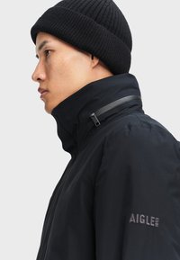 Giacca nera con colletto alto, design minimale e berretto a maglia a coste. Presenta un dettaglio con zip vicino al colletto e un logo sulla manica.