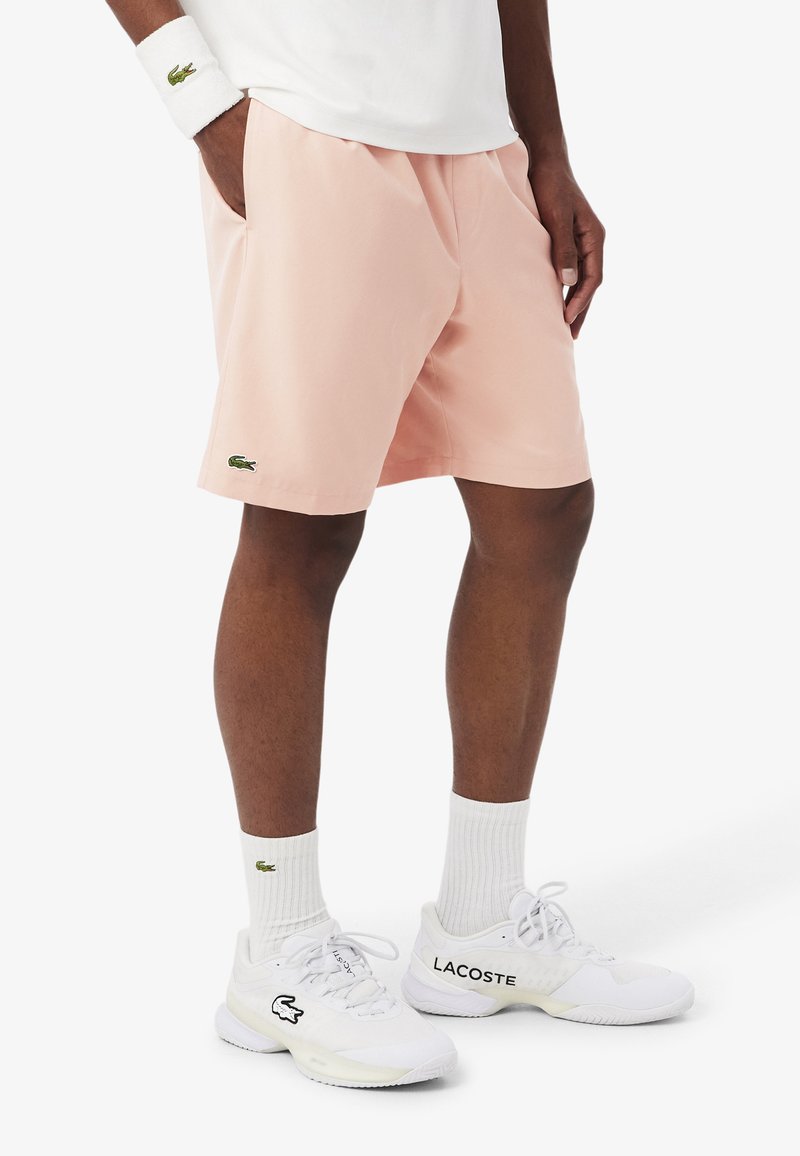 Person trägt hellrosa Shorts, weiße Socken, weiße Lacoste-Sneaker und ein weißes Armband und steht mit den Händen in den Taschen.