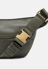 Olivegrünes Lederhandtasche mit verstellbarem Trageriemen und einer goldfarbenen Metallschnalle mit eingraviertem Branding. Glatte Textur, kompaktes Design.