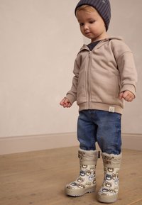 Sweat à capuche beige clair à fermeture éclair, t-shirt bleu foncé, jean en denim et bottes de pluie à motifs avec des visages d'animaux en beige, marron et gris.