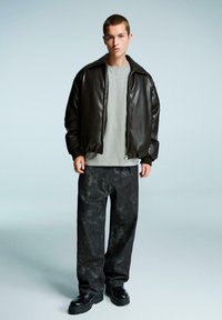 Giacca bomber in pelle marrone, t-shirt grigia, pantaloni scuri a fantasia e stivali neri. La giacca ha una chiusura con zip e polsini a costine.