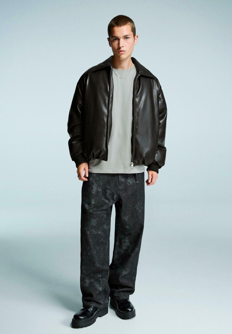 Giacca bomber in pelle marrone, t-shirt grigia, pantaloni scuri a fantasia e stivali neri. La giacca ha una chiusura con zip e polsini a costine.