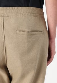 Pantalones beige con una textura sutil, que presentan un bolsillo trasero cosido y una cinturilla elástica. La tela parece duradera y suave.