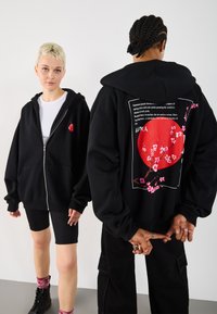 To modeller har på seg svarte hettegensere; foran er det en liten rød ferskenblomst, mens baksiden har en stor rød sirkel med rosa blomstergrener og tekst.
