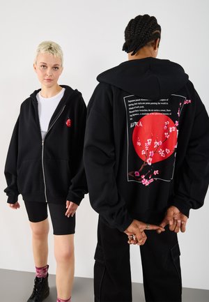 Dwie modelki mają na sobie czarne bluzy z kapturem; z przodu znajduje się mały czerwony kwiat brzoskwini, natomiast z tyłu duże czerwone koło z różowymi gałązkami kwiatów i napisem.
