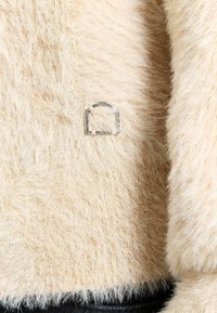 Tessuto fuzzy color crema con una consistenza morbida, caratterizzato da un accento logo argentato sul lato sinistro, su una base scura a contrasto.