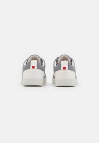 Sapatilhas cinzentas com sola e detalhe do calcanhar brancos, apresentando uma faixa vertical vermelha na parte de trás. Superior em tecido com um acabamento texturizado.