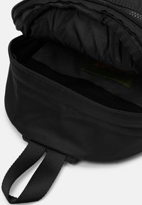 Zaino nero con tessuto testurizzato, chiusura con zip e due tracolle intrecciate. L'interno presenta un rivestimento liscia e un'etichetta verde.