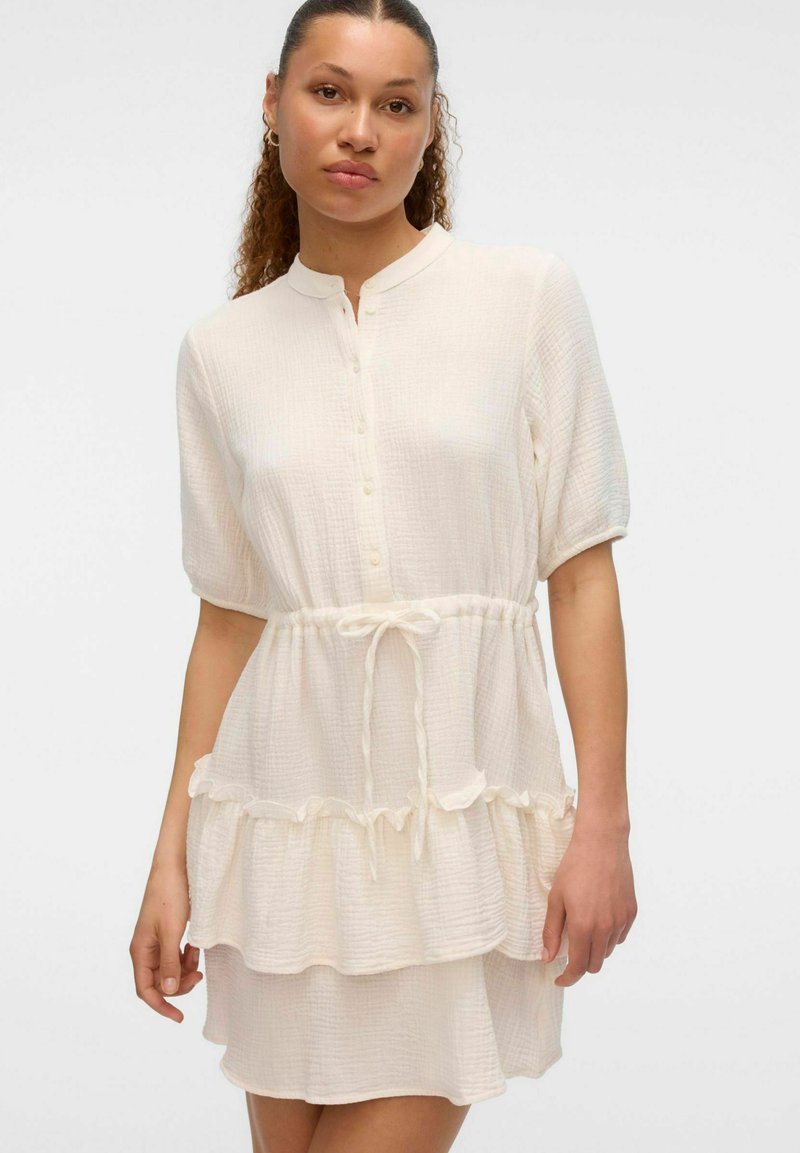 Robe blanche à volants en tissu texturé, avec un col boutonné, des manches courtes et une taille à cordon pour une silhouette ajustée.