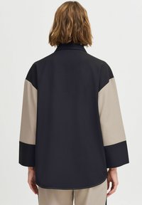 Cămașă cu mâneci lungi, având un design în blocuri de culoare neagră și bej, confecționată dintr-un material neted, cu guler și croială lejeră. Detalii de cusătură vizibile.