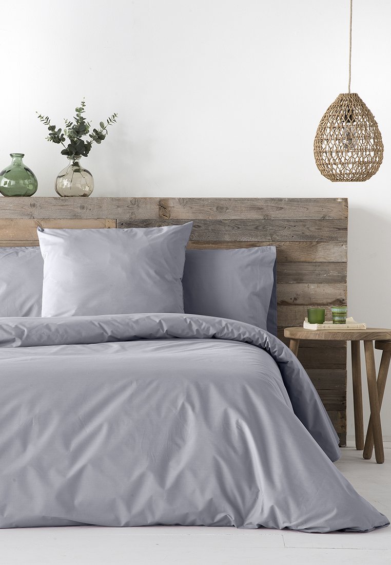 Set di biancheria da letto grigia con una trama di tessuto liscia, include due cuscini e un copriletto, abbinato a una testata in legno e oggetti decorativi.