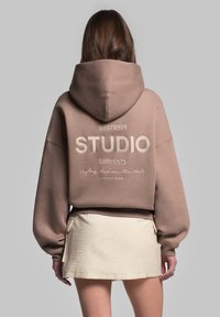 Ruskea yliystävällinen huppari, jossa on etulogo vaaleassa kermanvärissä, varustettuna naruhupulla ja ribbed-hihansuilla, yhdistettynä vaaleanbeigeen minihelmaan.